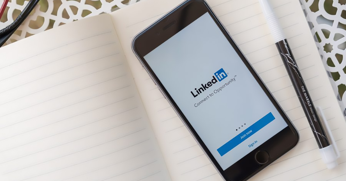 Smartphone displaying the LinkedIn login screen with the tagline &ldquo;Connect to Opportunity,&rdquo; placed on an open lined notebook beside a black-and-white pen.