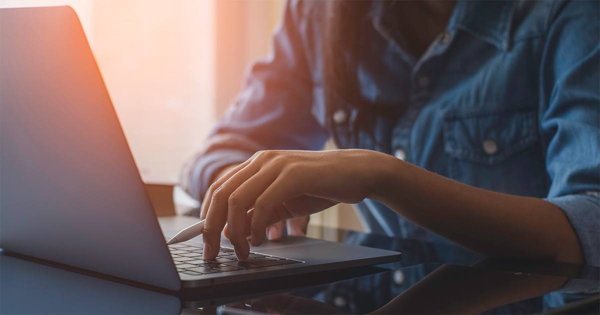 A woman who is casually dressed, working on a laptop, with one hand on the keyboard and warm sunlight streaming in from the left side of the frame.